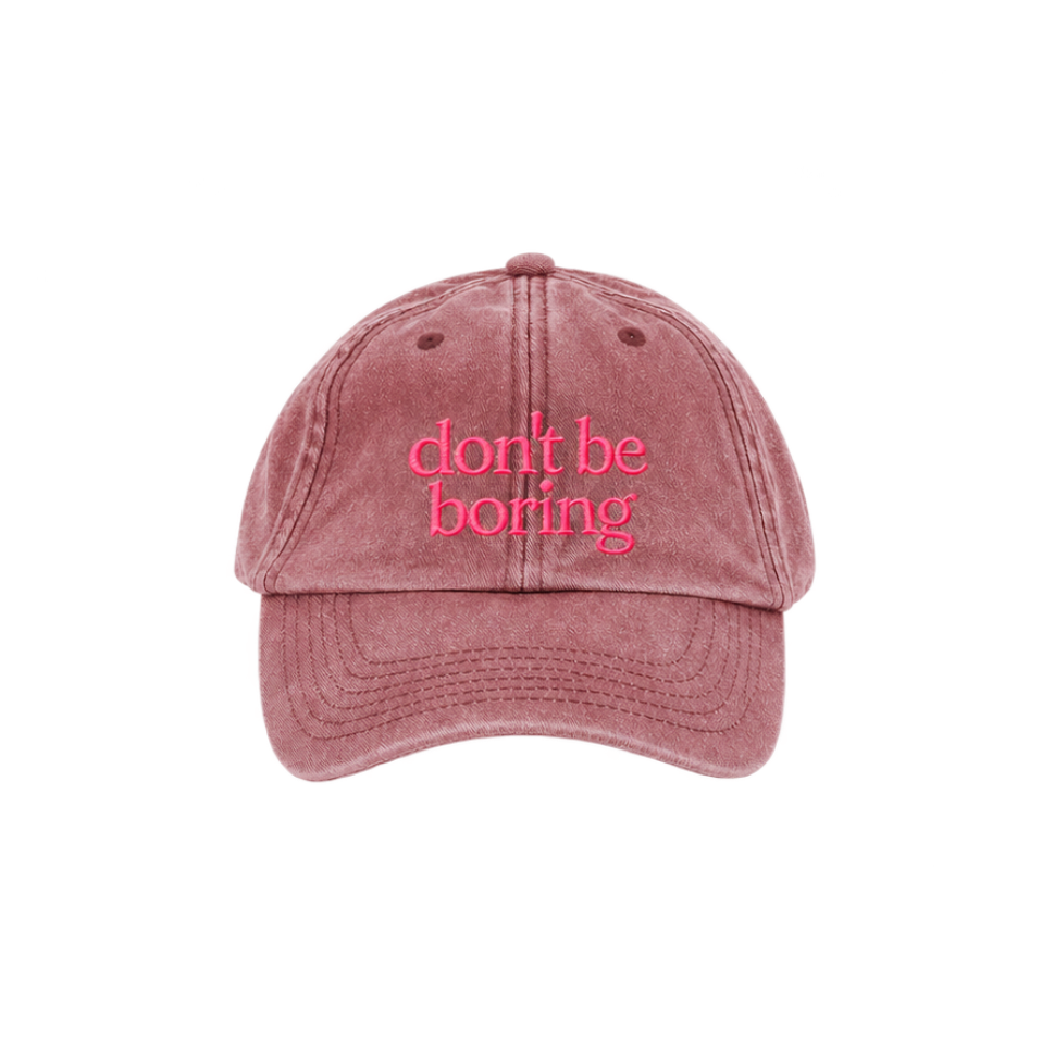 A washed dusty rose baseball cap with the phrase 'don't be boring' embroidered in pink script across the front, shown against a plain white background.